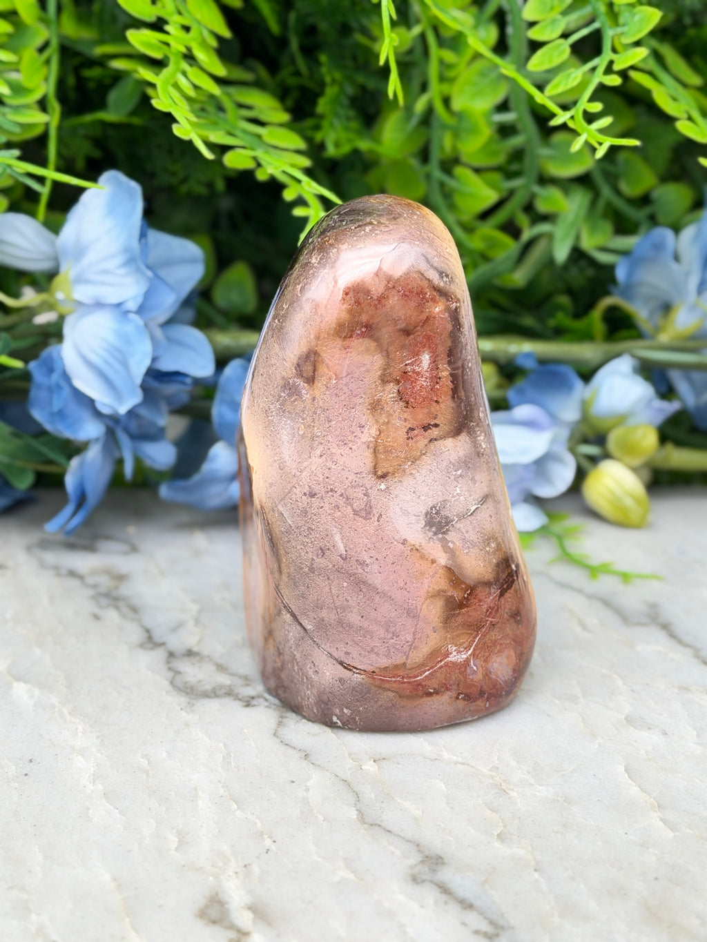 a large, irregularly shaped rock with a reddish-brown hue, placed on a marble surface. The rock is surrounded by a variety of blue flowers, creating a visually striking contrast.