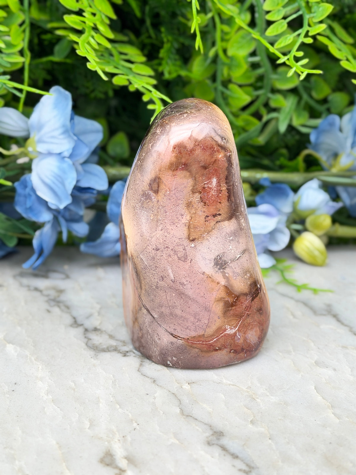 a large, irregularly shaped rock with a reddish-brown hue, placed on a marble surface. The rock is surrounded by a variety of blue flowers, creating a visually striking contrast.