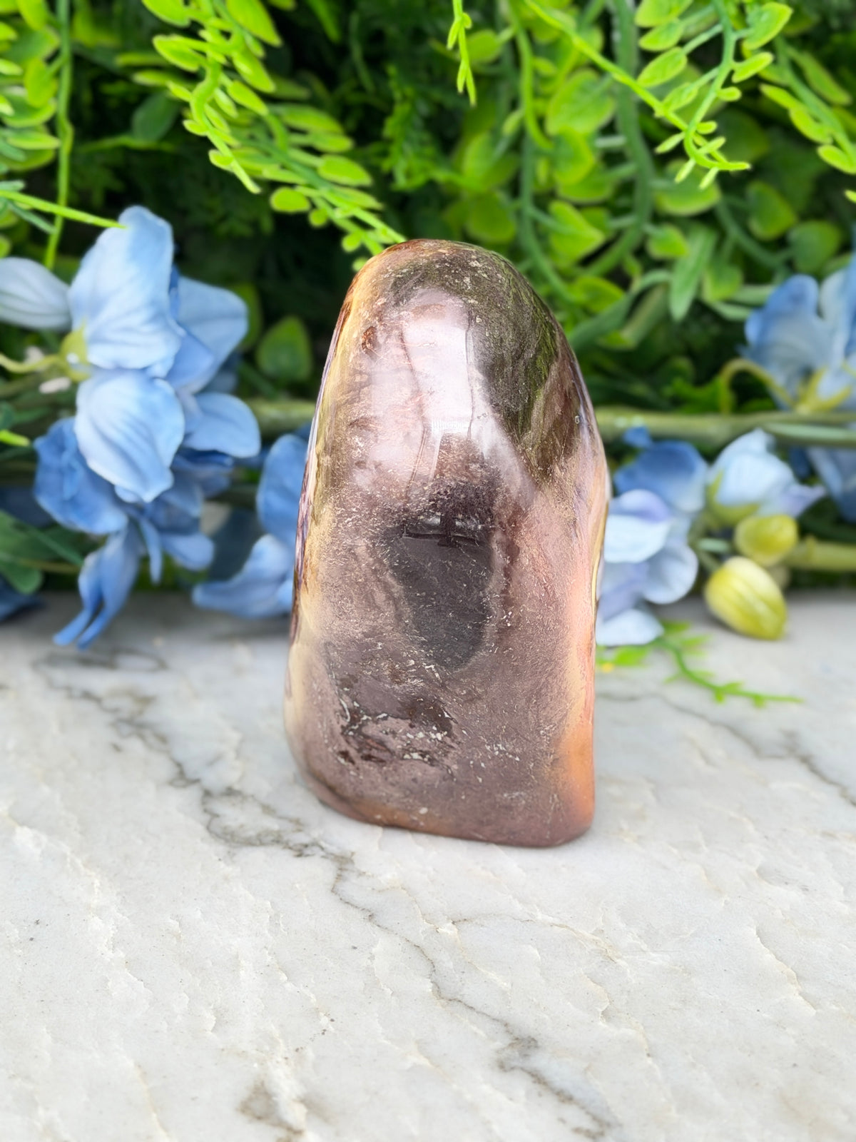 a brown, irregularly shaped rock with a dark, speckled surface, placed on a white marble surface. The rock is surrounded by blue flowers, creating a visually appealing contrast between the natural and the artificial elements.