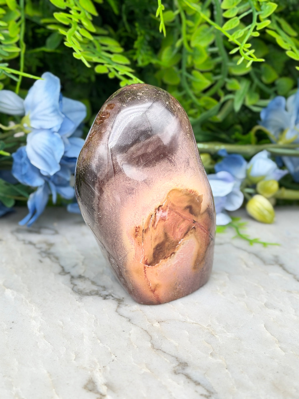 a jadeite, a type of agate, with a unique brownish-red hue and a prominent crack running through it, set against a backdrop of lush green foliage and delicate blue flowers.