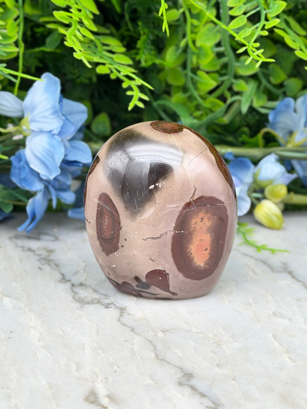 A brown and beige jadeite gemstone with a speckled pattern sits on a white marble surface, surrounded by blue flowers.