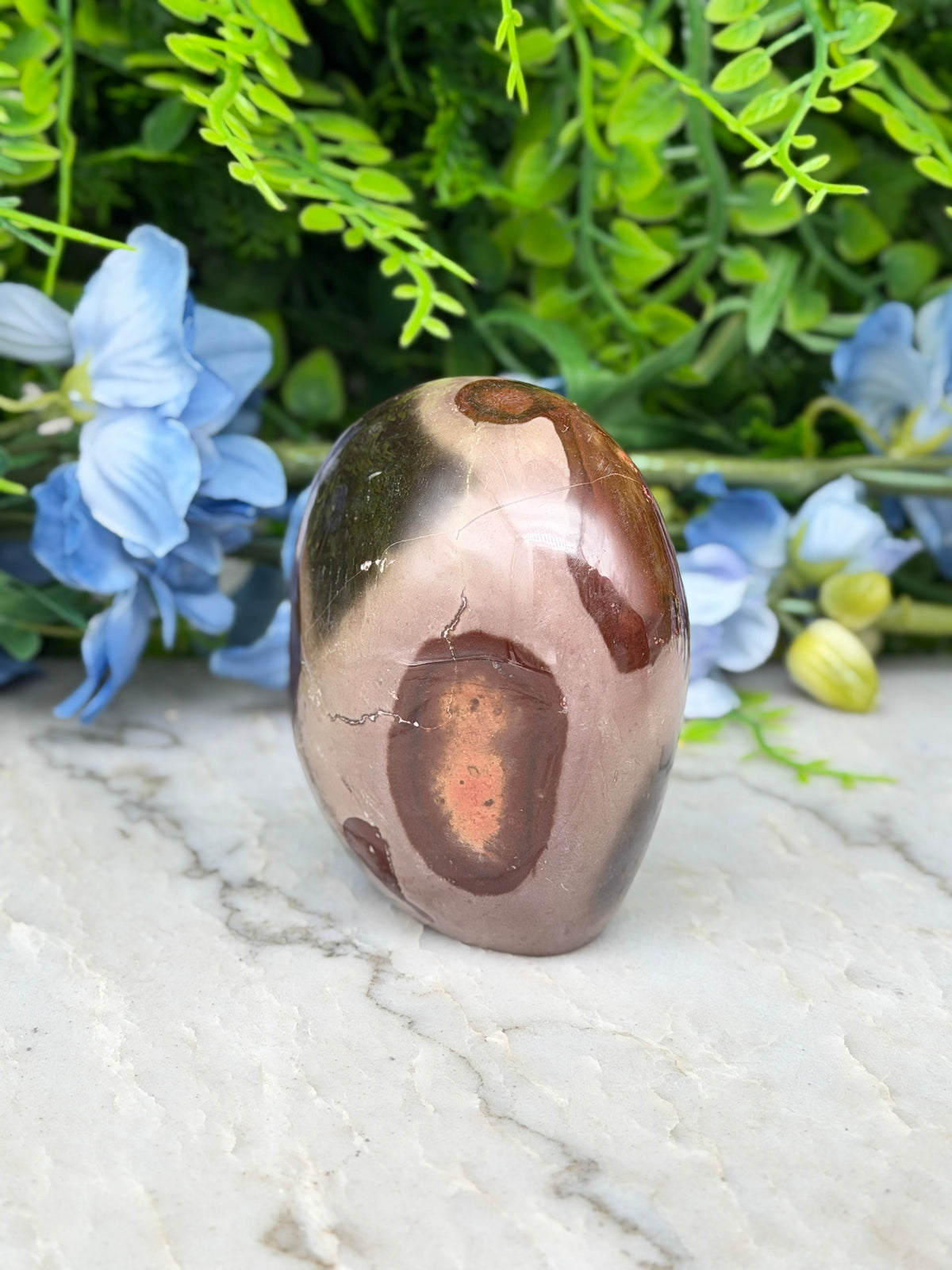 A brown jadeite gemstone with a unique pattern sits on a white marble surface, surrounded by blue flowers and green foliage.