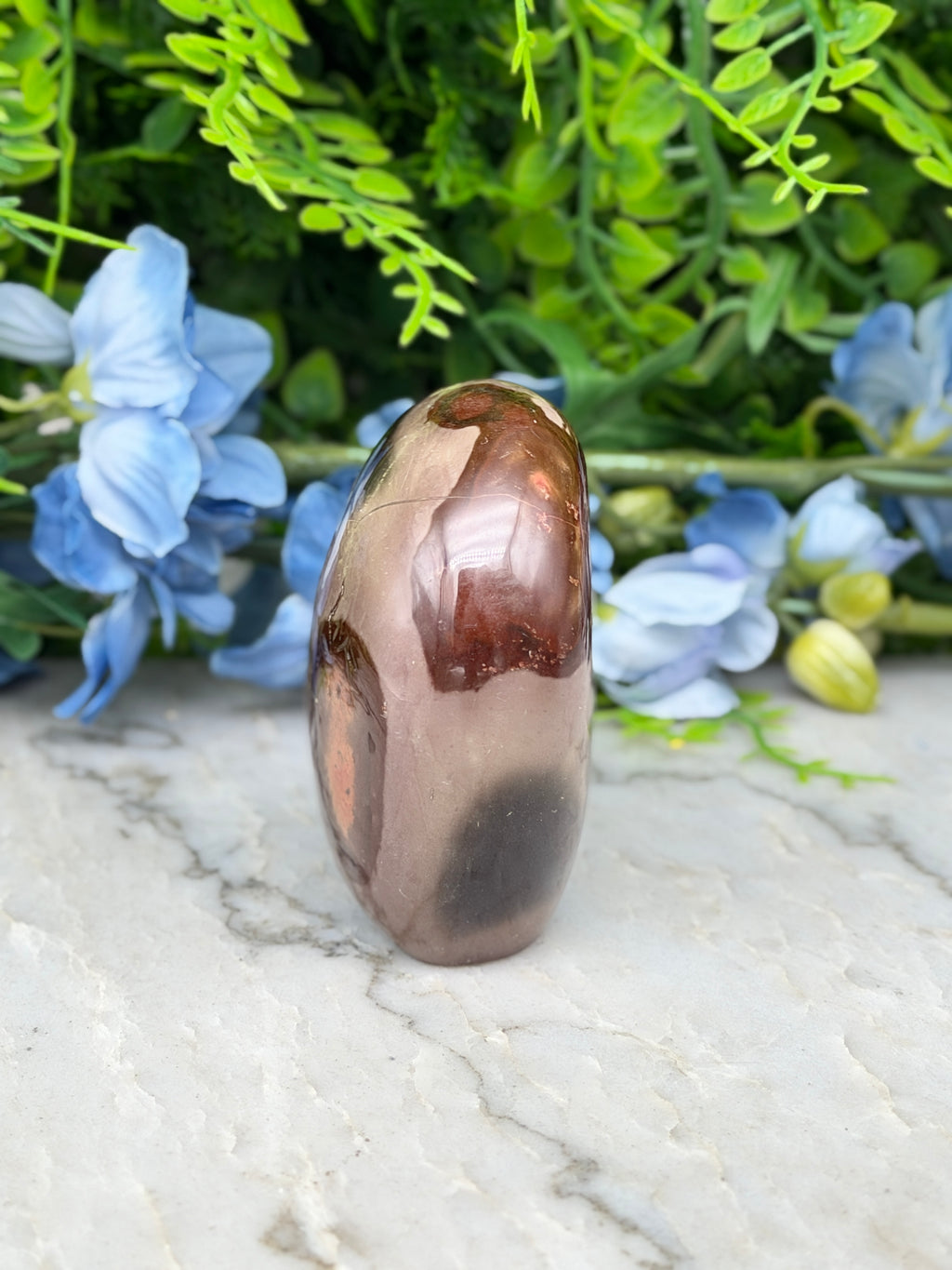a brown, oval-shaped stone or gemstone placed on a white marble surface, surrounded by blue flowers.