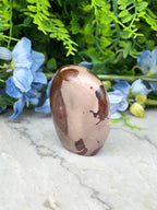 A brown and beige jadeite stone is placed on a marble surface, surrounded by blue flowers and green foliage.