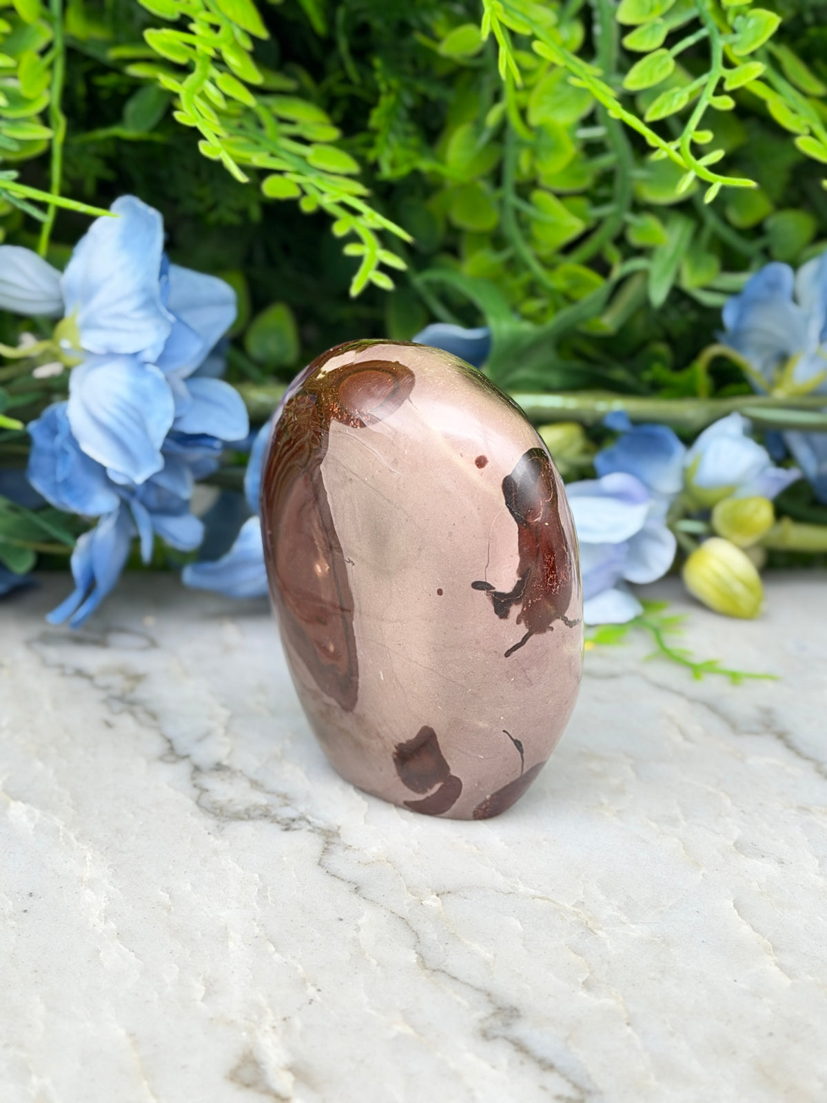 A brown and beige jadeite stone is placed on a marble surface, surrounded by blue flowers and green foliage.