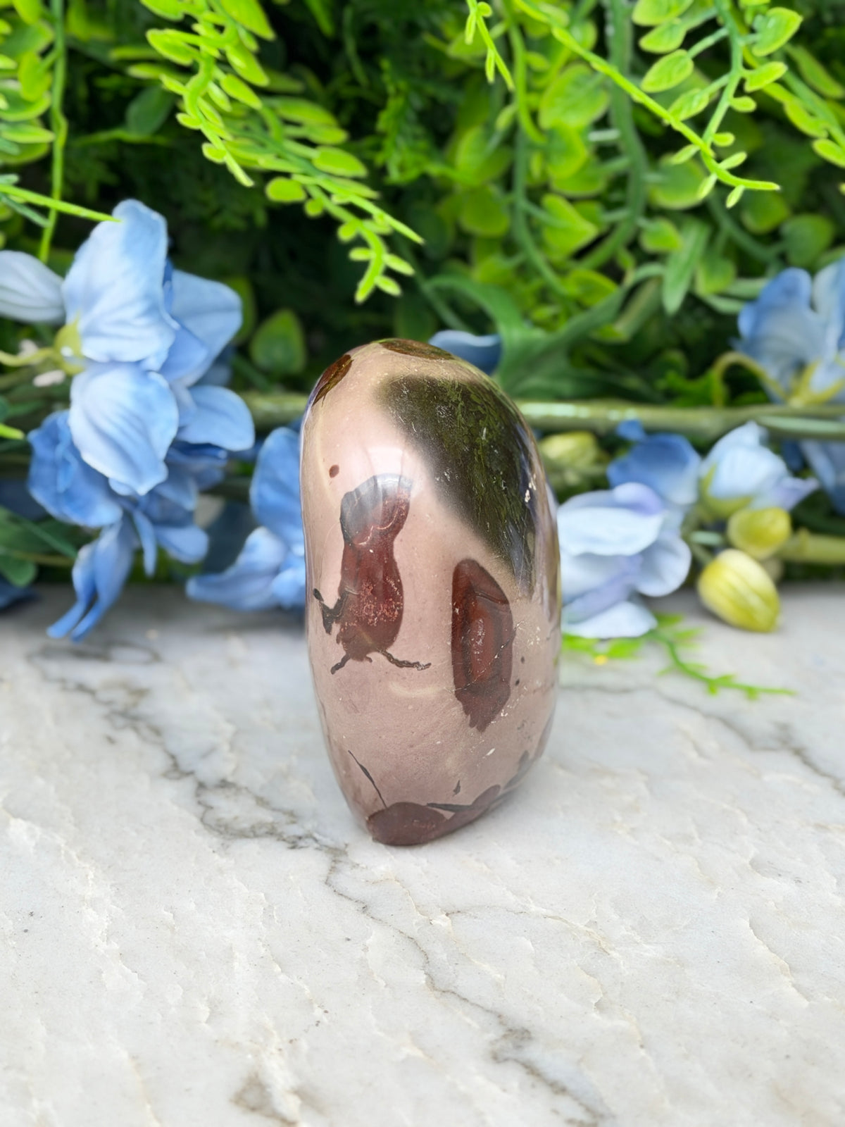 A jadeite gemstone with a brownish hue and a black pattern is displayed on a marble surface, surrounded by blue flowers.