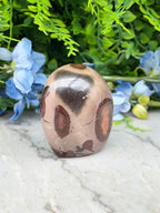 A brown and black speckled jadeite gemstone is displayed on a white marble surface, surrounded by blue flowers.
