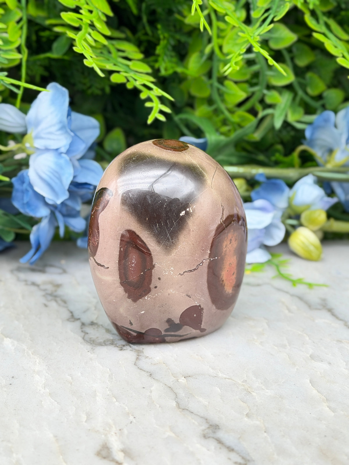 A brown and black speckled jadeite gemstone is displayed on a white marble surface, surrounded by blue flowers.