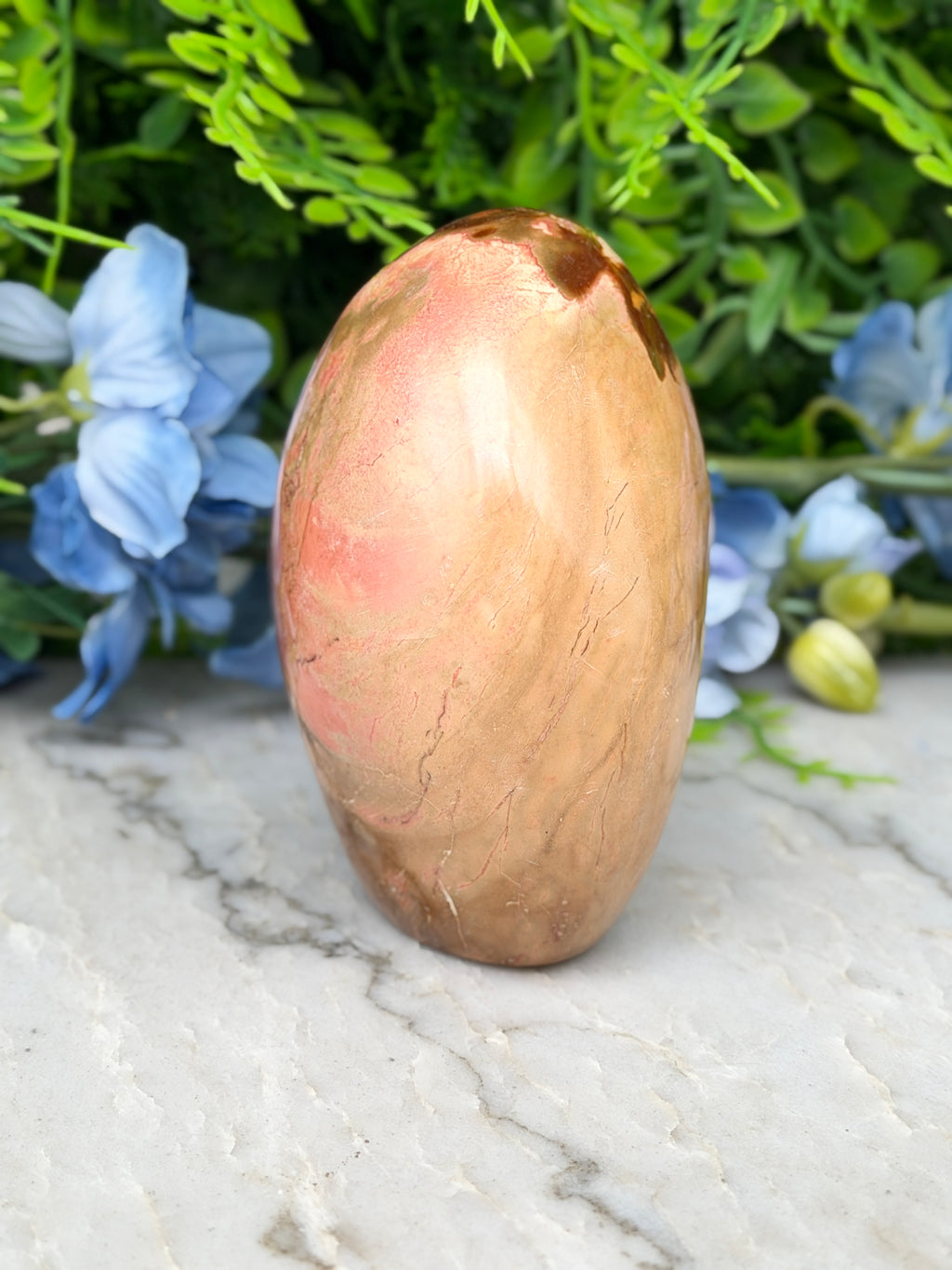 a brown, oval-shaped stone or rock with a reddish-brown hue, placed on a white marble surface. The stone is surrounded by a variety of blue and green flowers, creating a visually appealing contrast.