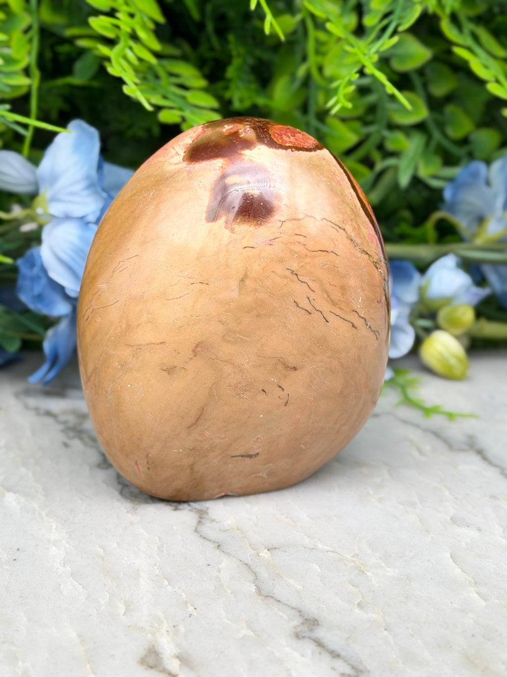 a brown, oval-shaped object with a reddish-brown hue, placed on a white surface. The object appears to be a piece of wood or a stone, with a natural, earthy texture. In the background, there are blue flowers, adding a touch of color and life to the scene.