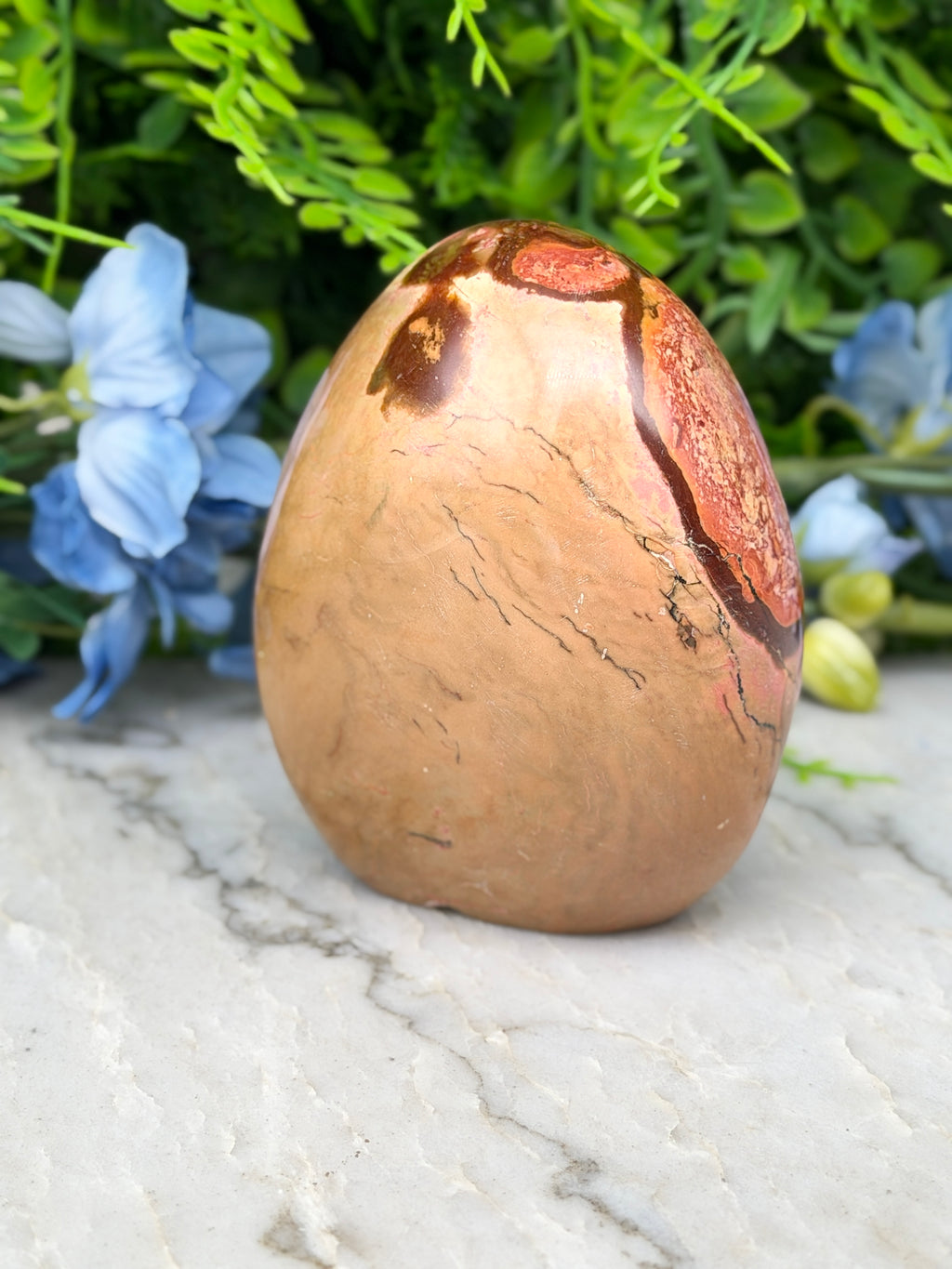 a brown, speckled rock with a striking pattern, placed on a marble surface. The rock is surrounded by a variety of blue flowers, creating a visually appealing contrast.