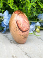 a jadeite, a type of jadeite, with a distinctive pinkish hue and a rough, textured surface. The jadeite is placed on a marble surface, surrounded by blue flowers, creating a visually appealing contrast between the natural and the artificial elements.