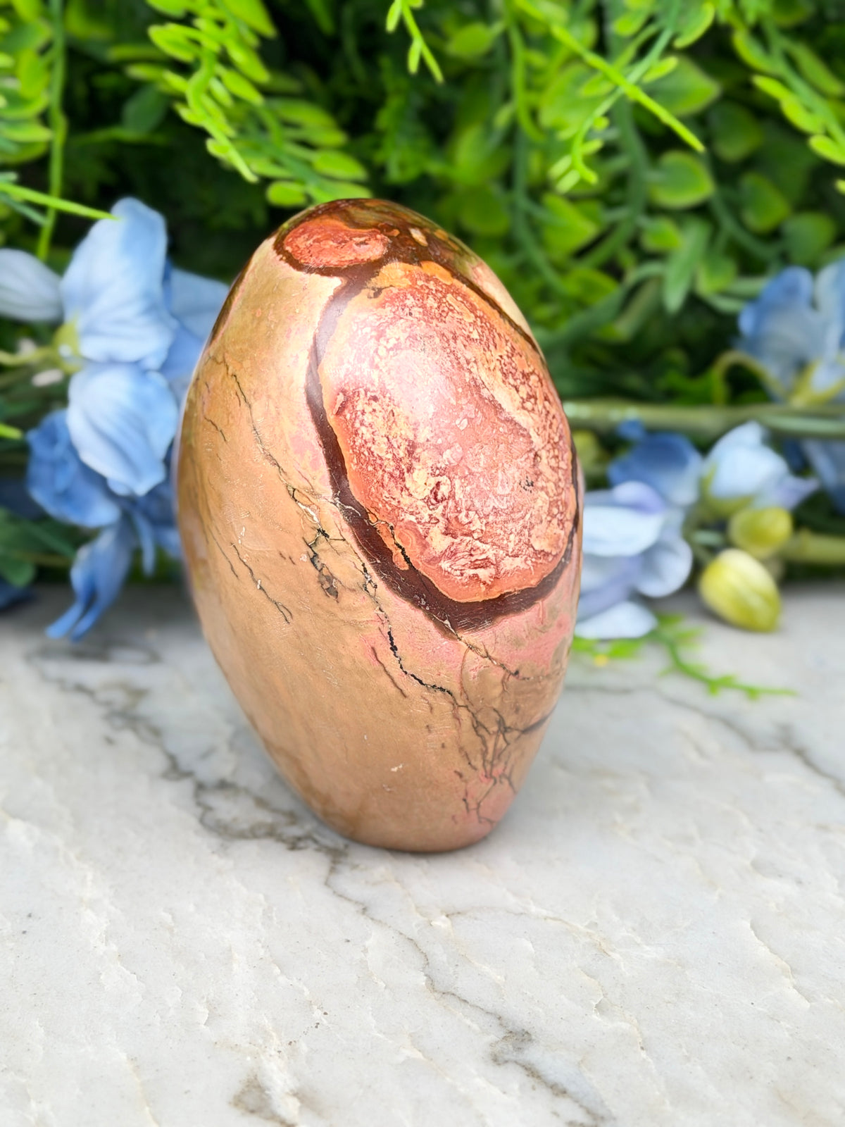 a jadeite, a type of jadeite, with a distinctive pinkish hue and a rough, textured surface. The jadeite is placed on a marble surface, surrounded by blue flowers, creating a visually appealing contrast between the natural and the artificial elements.
