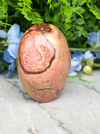 A pinkish-brown jadeite gemstone with a distinctive pattern sits on a white marble surface, surrounded by blue flowers.