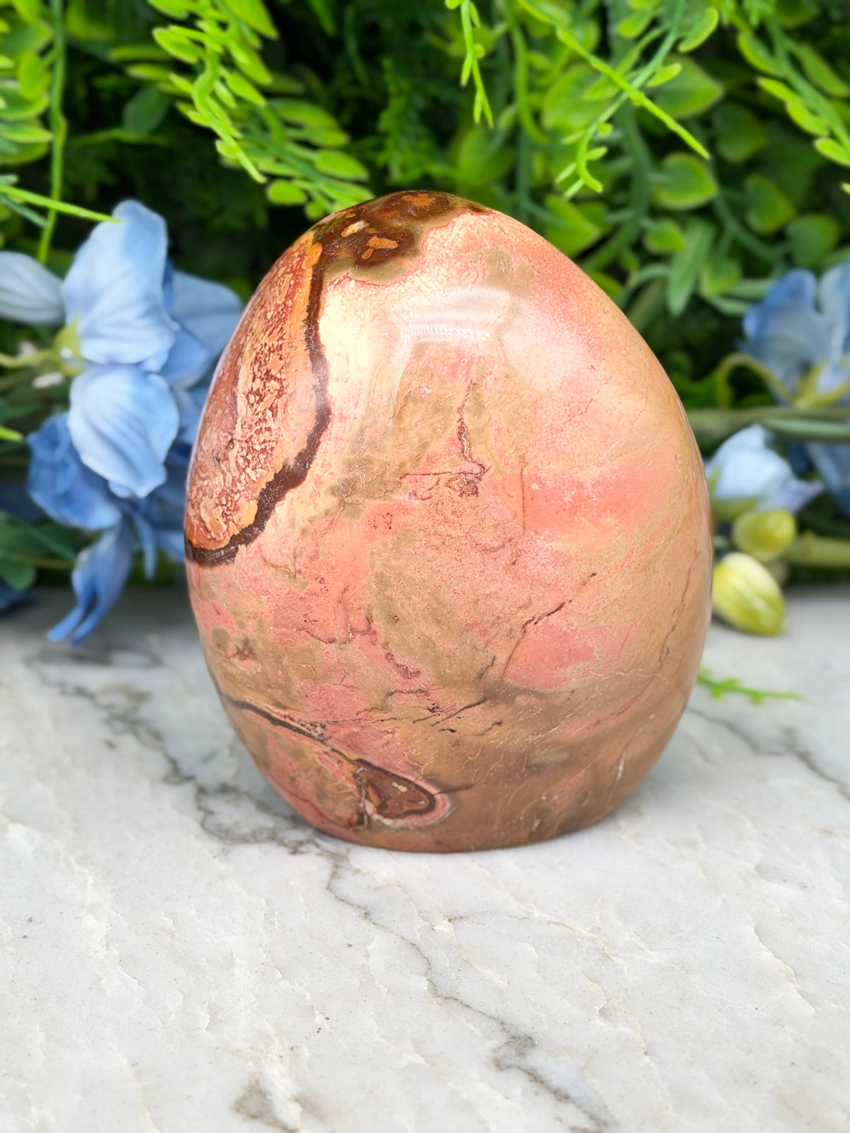 A pinkish-brown jadeite rock with a unique pattern sits on a marble surface, surrounded by blue flowers.