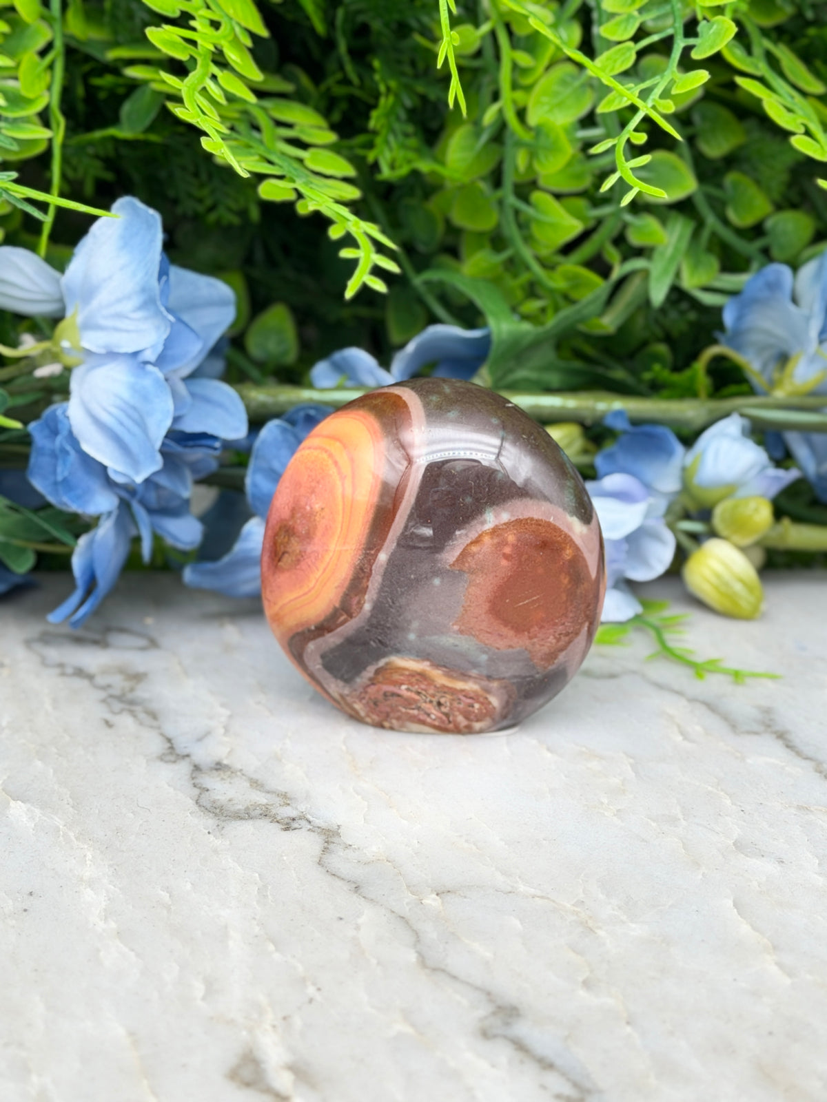 a large, round, brown and orange stone, possibly an agate, placed on a marble surface surrounded by blue flowers.