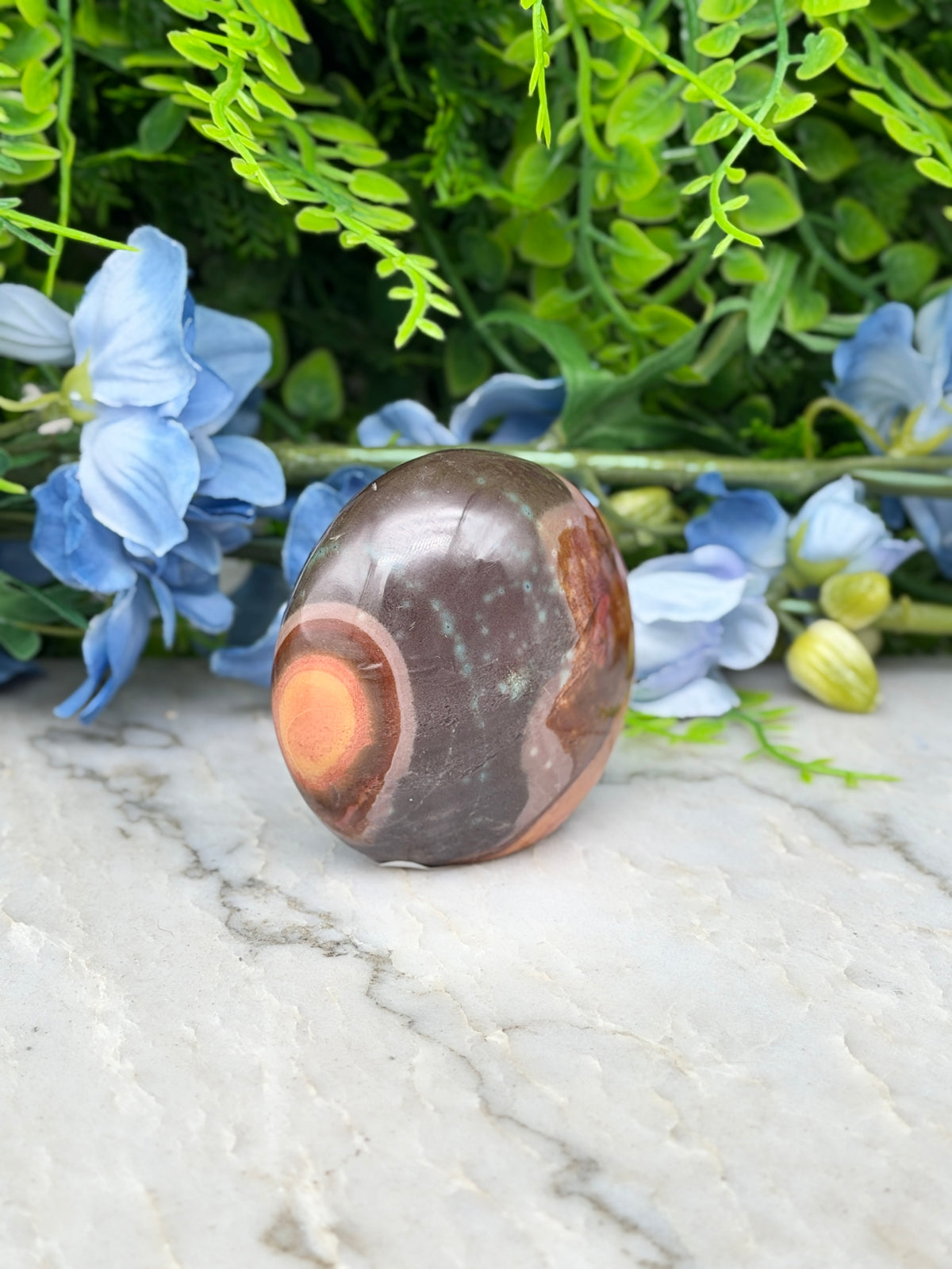 a brown and orange rock with a circular pattern, placed on a white marble surface. The rock is surrounded by blue flowers and green foliage, creating a visually appealing and natural setting.