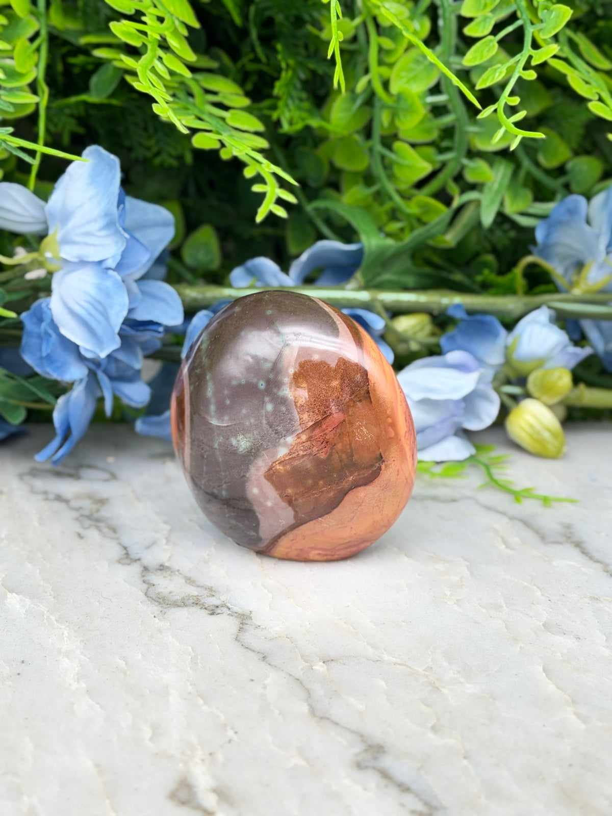 a brown and orange stone, possibly a jade or amber, placed on a white marble surface. The stone is surrounded by blue flowers and green foliage, creating a visually appealing and harmonious composition.