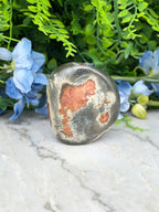 a large, round, grey rock with a reddish-brown hue, placed on a white marble surface. The rock is surrounded by a variety of blue and green flowers, creating a visually appealing contrast between the natural and man-made elements.
