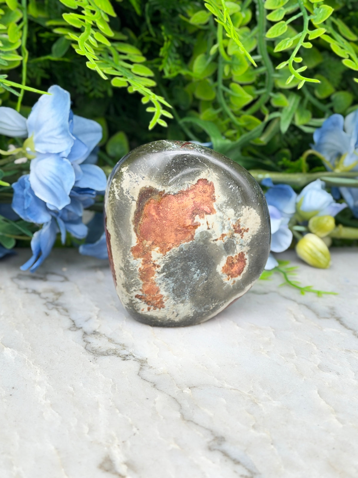 a large, round, grey rock with a reddish-brown hue, placed on a white marble surface. The rock is surrounded by a variety of blue and green flowers, creating a visually appealing contrast between the natural and man-made elements.