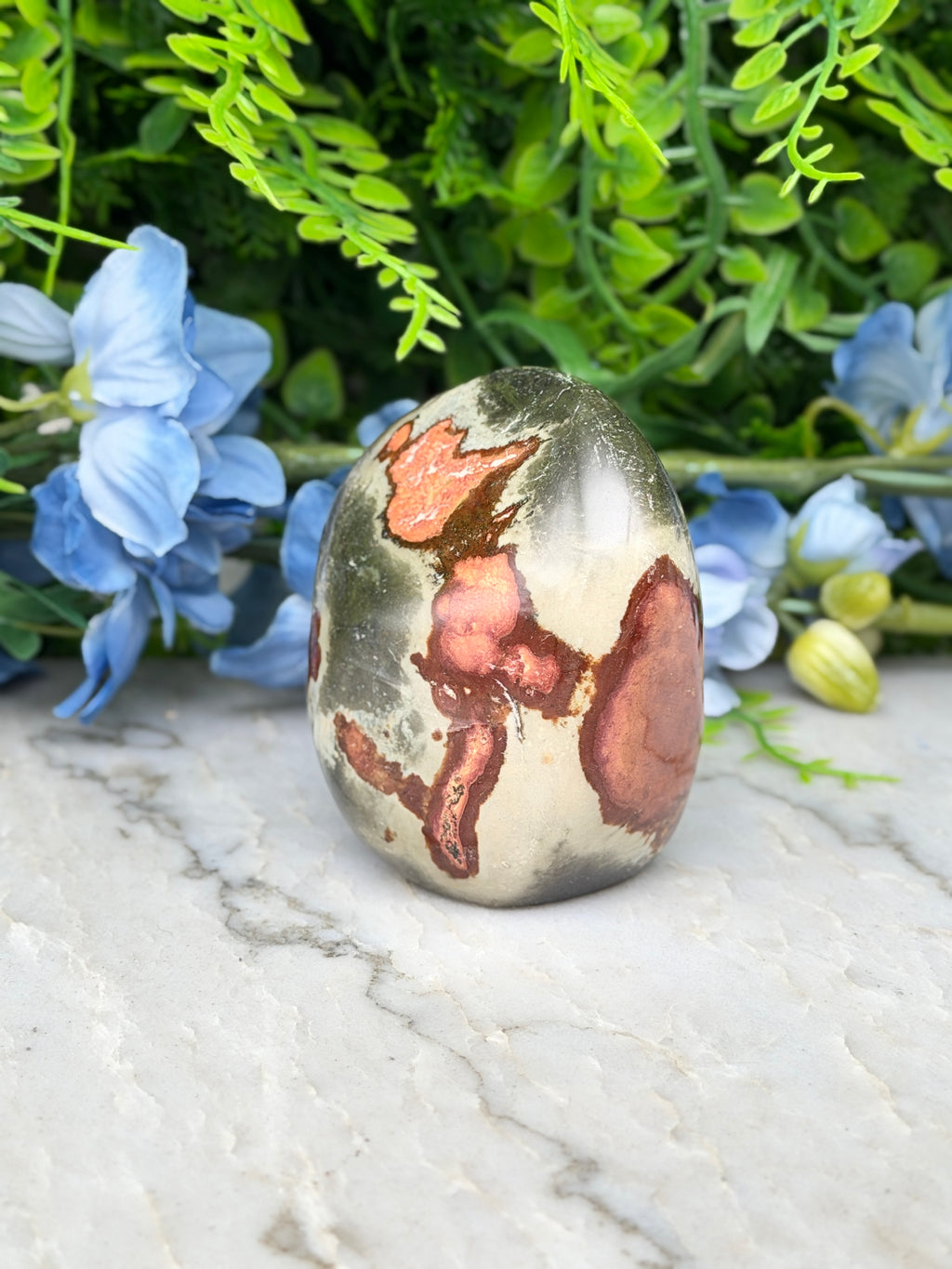 a jade egg, a natural stone with a unique, colorful pattern, placed on a marble surface surrounded by blue flowers.