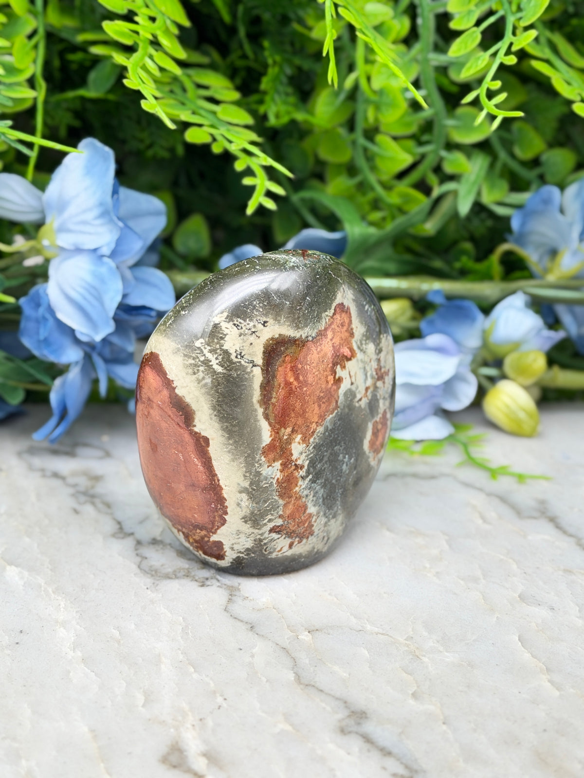 a large, round, and colorful stone with a unique pattern, placed on a marble surface. The stone has a mix of blue and brown hues, and it appears to be a natural formation. The stone is surrounded by a variety of green plants and flowers, creating a vibrant and visually appealing scene.