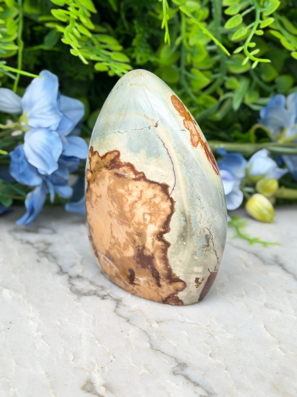 a unique, textured rock formation with a mix of brown and orange hues, set against a backdrop of lush green foliage and delicate blue flowers.