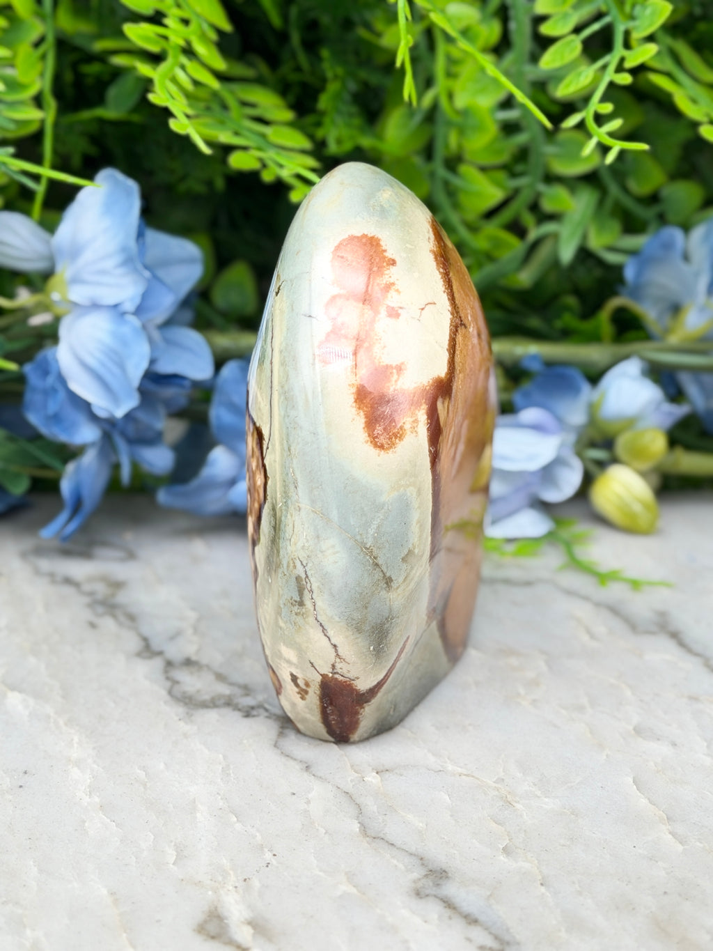 A jadeite, a type of agate, is displayed on a marble surface, surrounded by blue flowers.