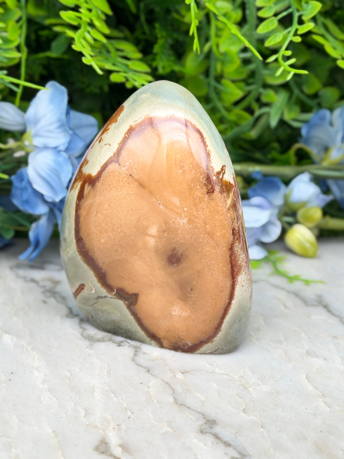 a jadeite, a type of agate, with a unique brownish-orange hue and a distinctive heart-shaped pattern, set against a backdrop of lush green foliage and delicate blue flowers.
