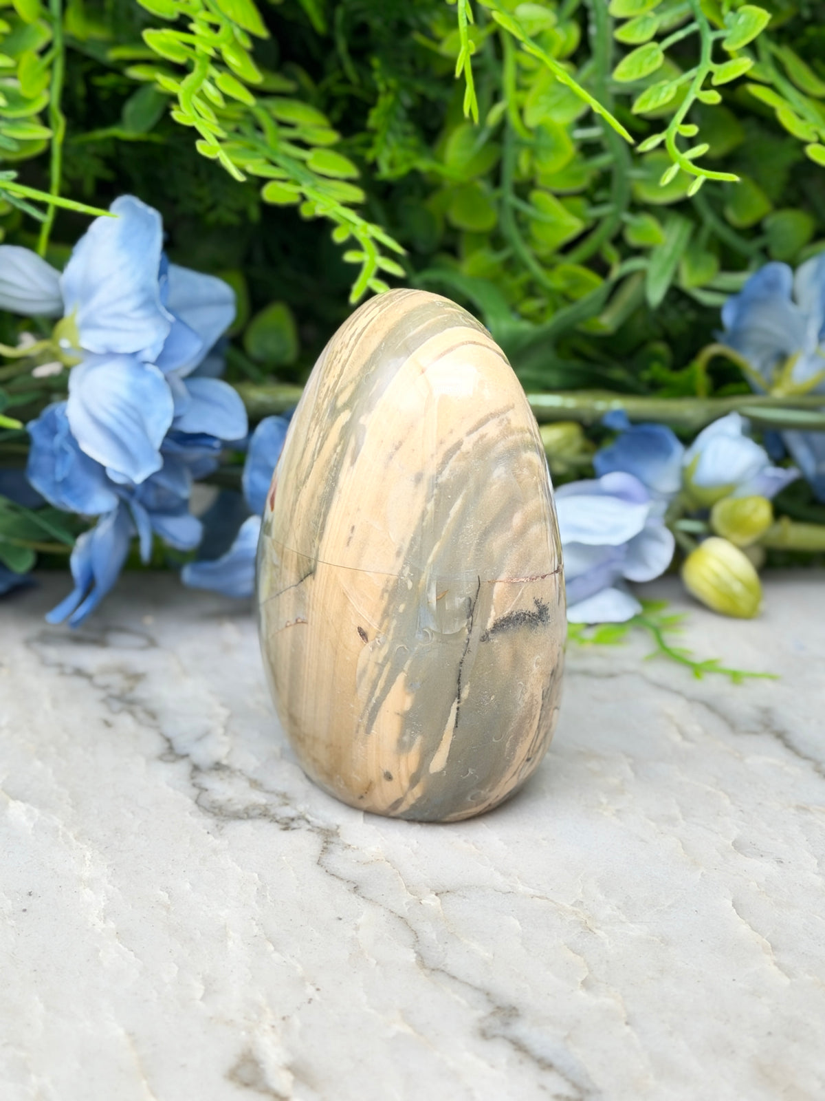 a marble-like stone or egg-shaped object, possibly a piece of jewelry or a decorative item, placed on a marble surface. The background is filled with lush green foliage, creating a natural and serene atmosphere.