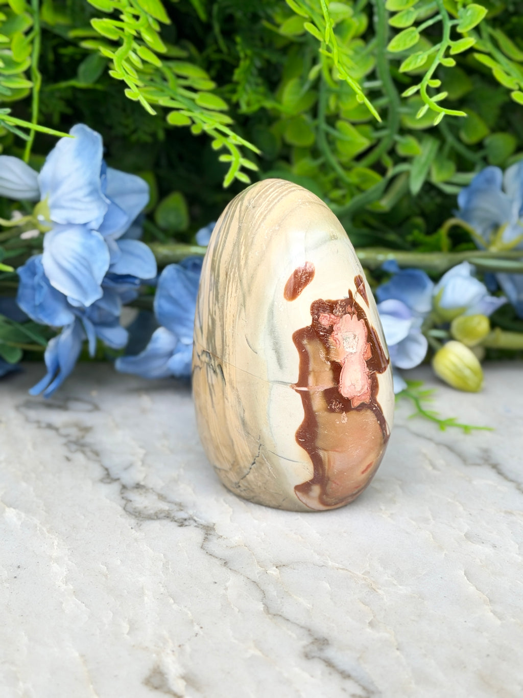 a natural stone, possibly an egg-shaped piece, placed on a marble surface. The stone has a unique pattern and is surrounded by blue flowers, creating a visually appealing contrast.
