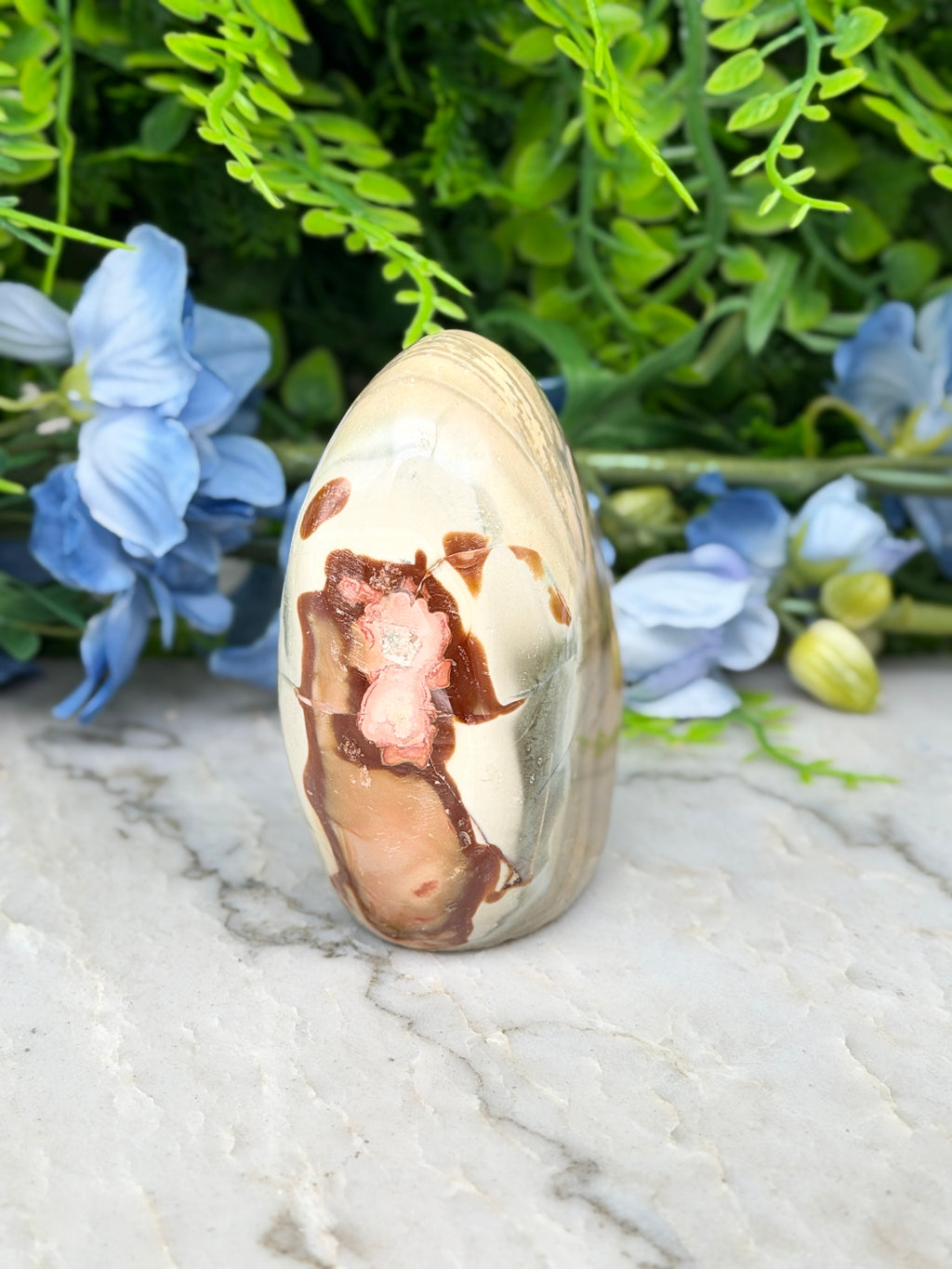 a jadeite gemstone with a unique pinkish hue, set against a backdrop of lush green foliage and delicate blue flowers.