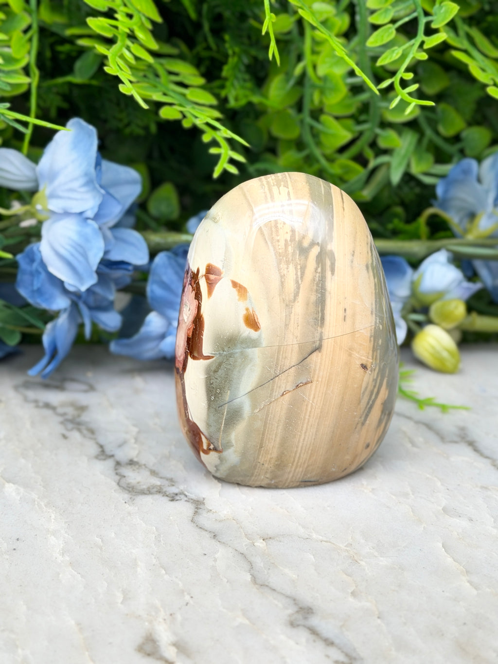 a jadeite, a type of jadeite gemstone, sitting on a marble surface. The jadeite has a unique pattern of light and dark colors, and it is surrounded by blue flowers and green foliage.