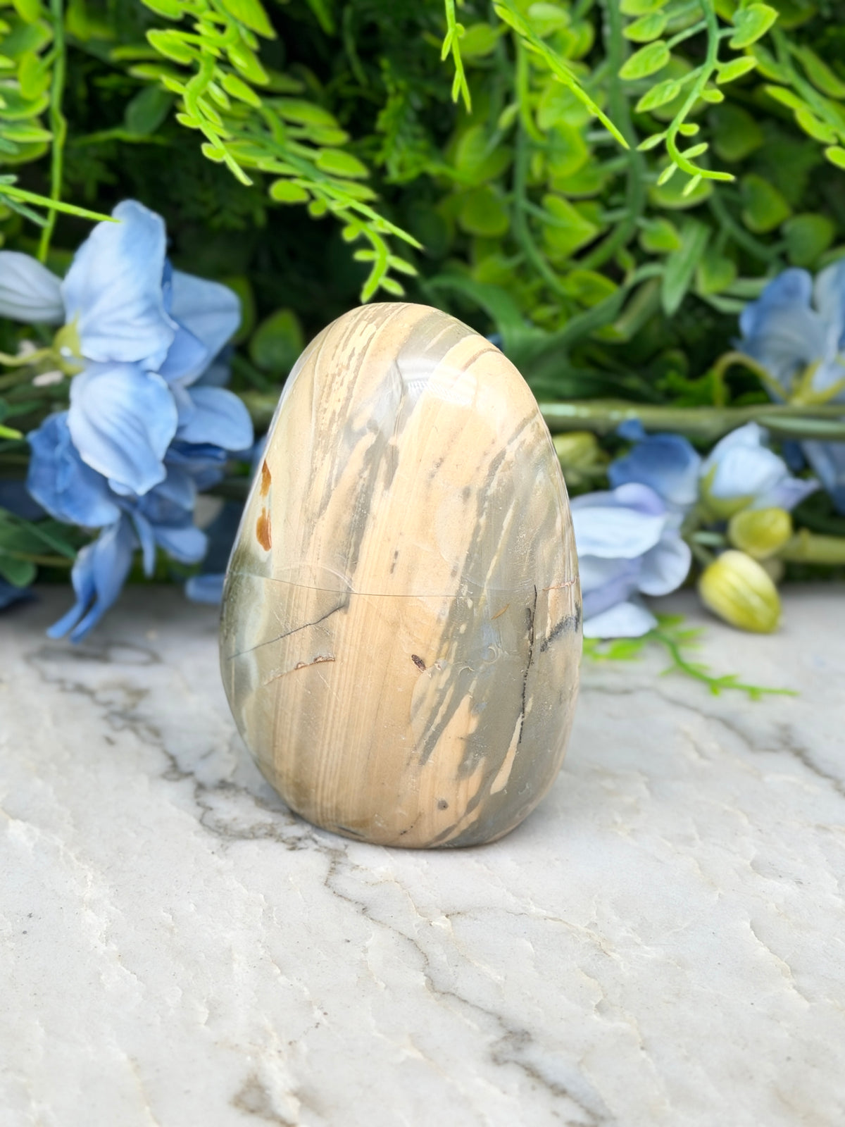 a natural stone, possibly a jade or slate rock, placed on a marble surface. The stone has a unique pattern and is surrounded by blue flowers, creating a visually appealing contrast.