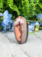 A brown and orange jadeite gemstone is displayed on a marble surface, surrounded by blue flowers.