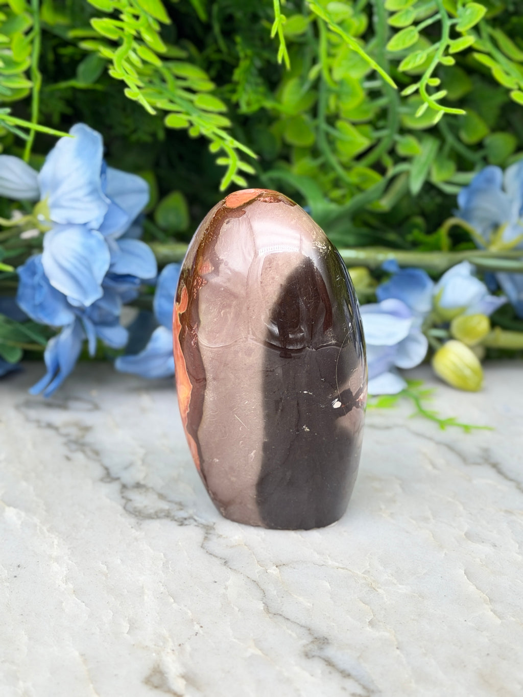 a brown and black stone, possibly a jade or amber, placed on a marble surface. The stone is surrounded by blue flowers, creating a visually appealing contrast.