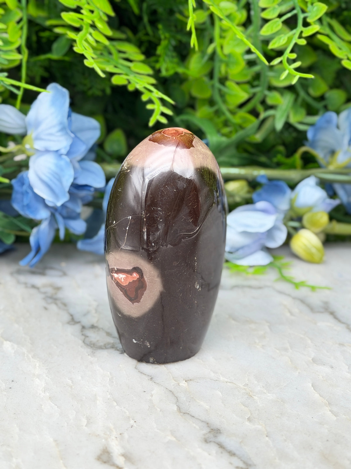 A dark brown, oval-shaped stone with a red spot on it, placed on a white surface with blue flowers in the background.