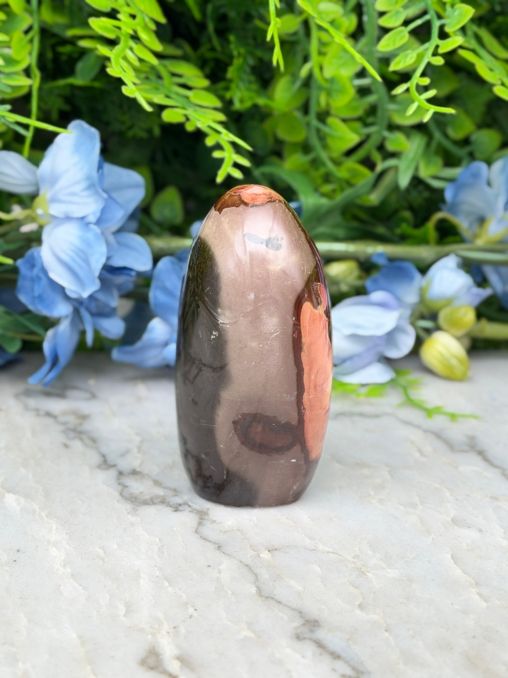 A brown and orange jadeite gemstone is displayed on a white marble surface, surrounded by blue flowers.