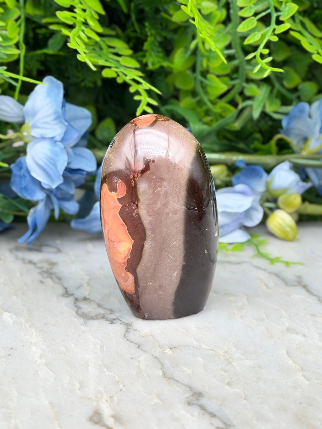 A brown and orange jadeite gemstone is displayed on a white marble surface, surrounded by blue flowers.