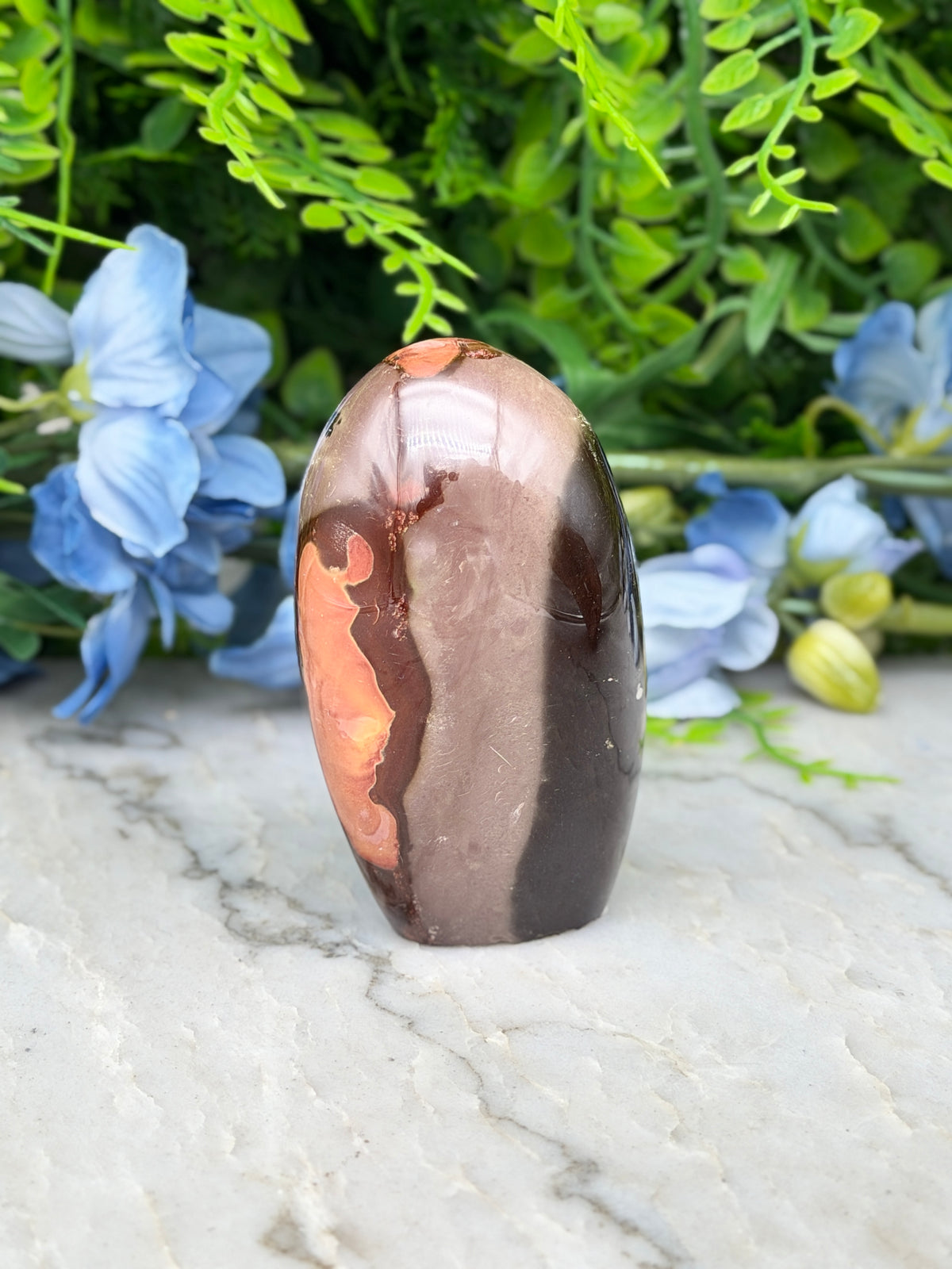 A brown and orange jadeite gemstone is displayed on a white marble surface, surrounded by blue flowers.