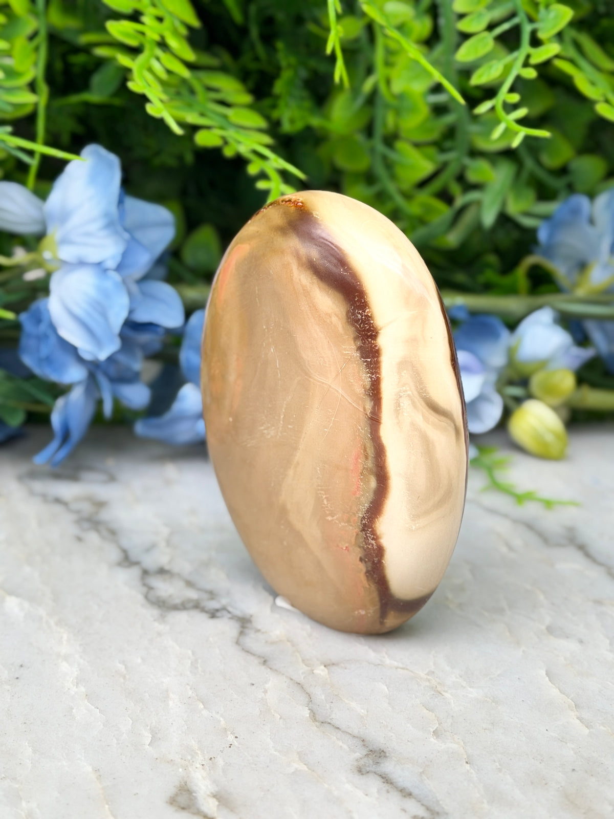 a large, oval-shaped stone with a brownish-brown hue, set against a backdrop of lush green foliage and delicate blue flowers.