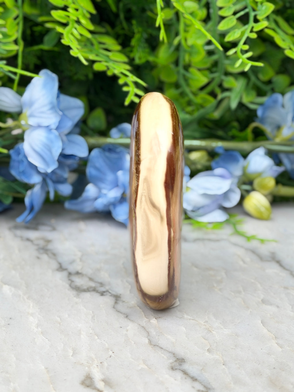 a tall, narrow, and brown-colored stone or gemstone, which appears to be a piece of amber or amberite, placed on a marble surface. The stone is surrounded by a variety of blue flowers, creating a visually appealing contrast between the natural and the artificial elements.
