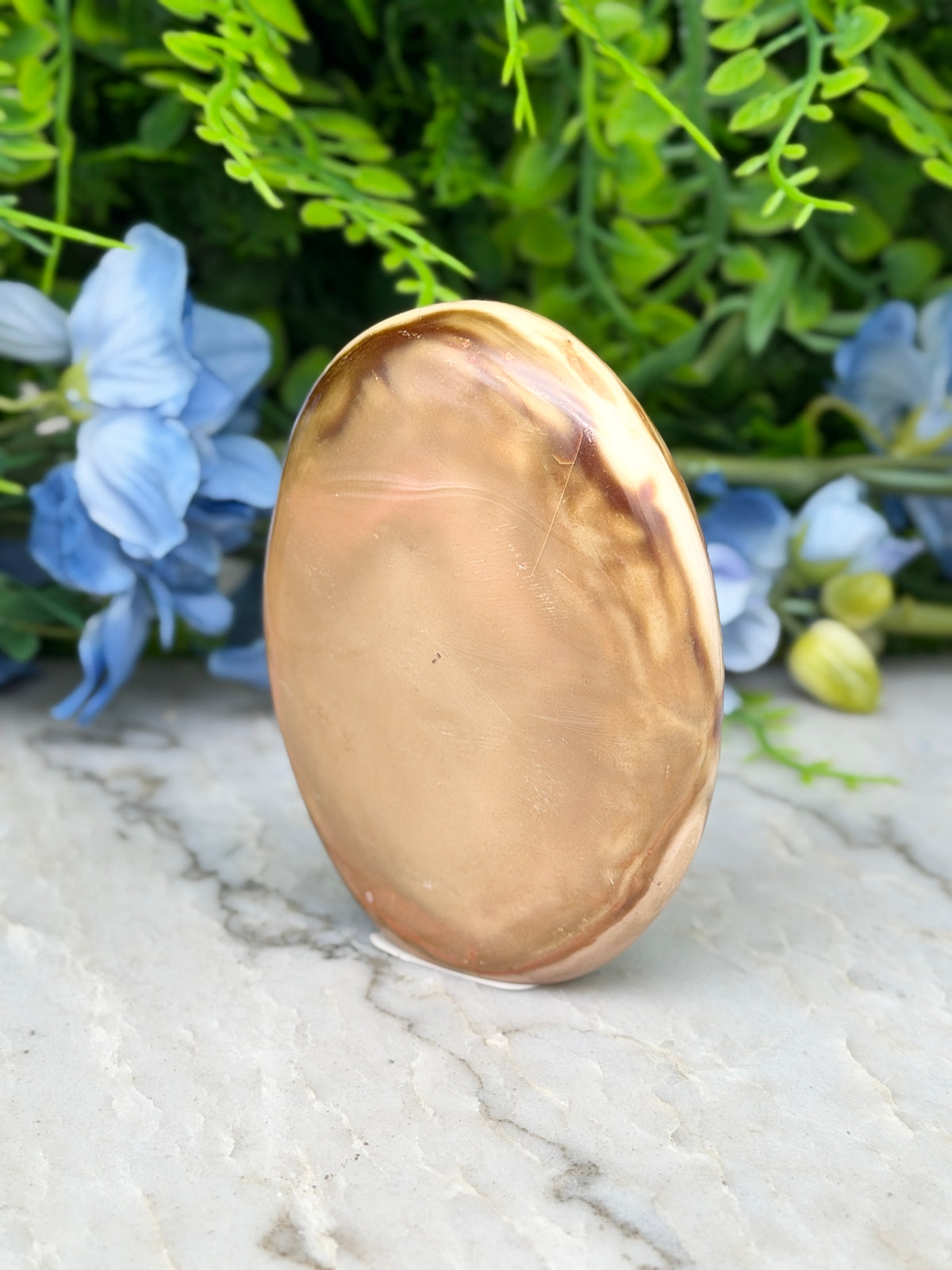 a large, oval-shaped stone or rock with a warm, golden hue, placed on a marble surface. The stone is surrounded by a variety of blue flowers, creating a visually appealing contrast.