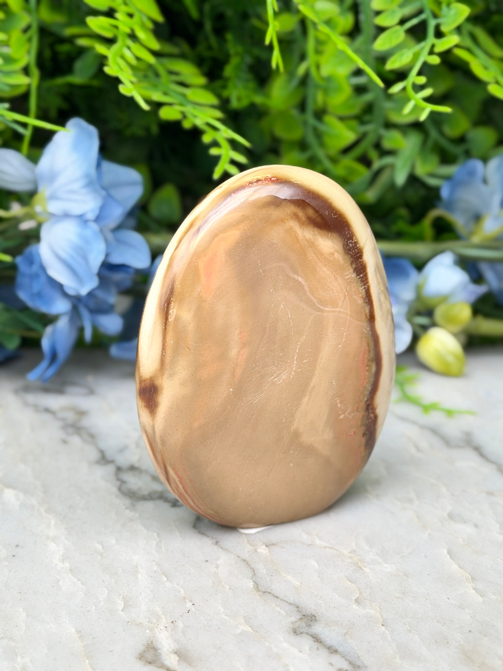 a brown oval-shaped stone or rock with a marbled pattern, placed on a white marble surface. The stone is surrounded by blue flowers, creating a visually appealing contrast.