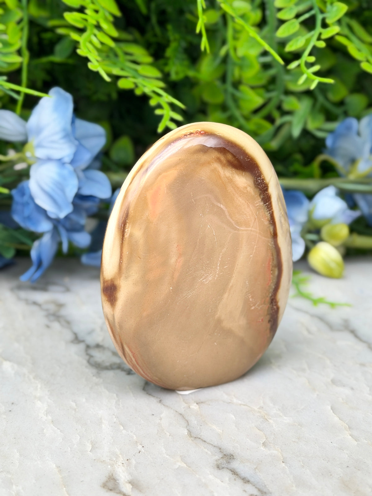 a brown oval-shaped stone or rock with a marbled pattern, placed on a white marble surface. The stone is surrounded by blue flowers, creating a visually appealing contrast.