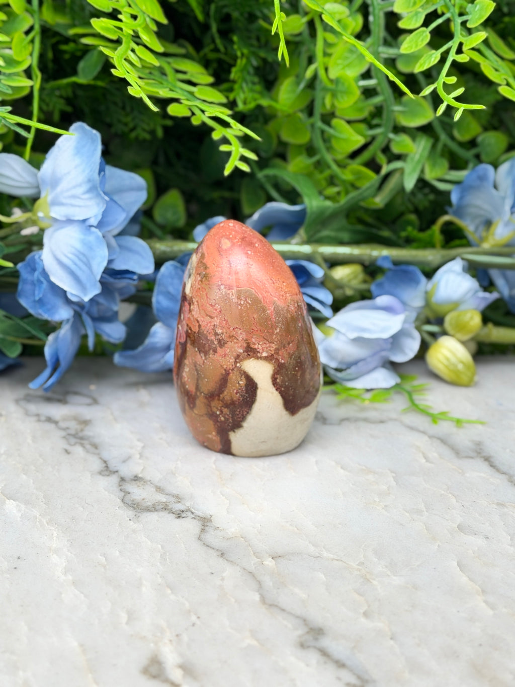 a red and brown speckled rock sitting on a marble surface, surrounded by blue flowers and green foliage.