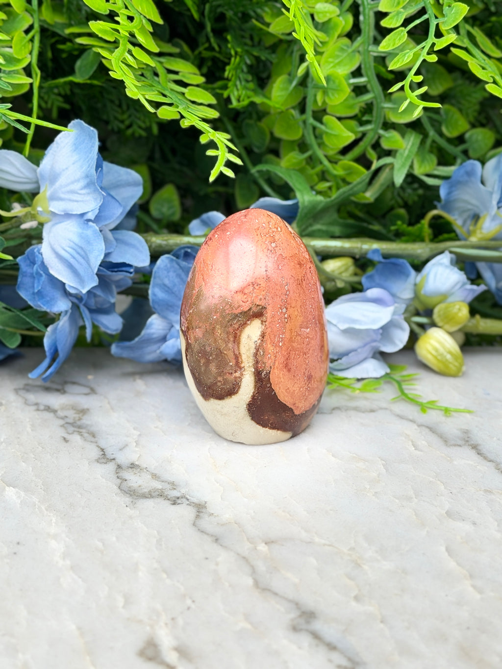 a single, vibrant orange and brown jade egg placed on a marble surface, surrounded by a lush display of blue flowers.