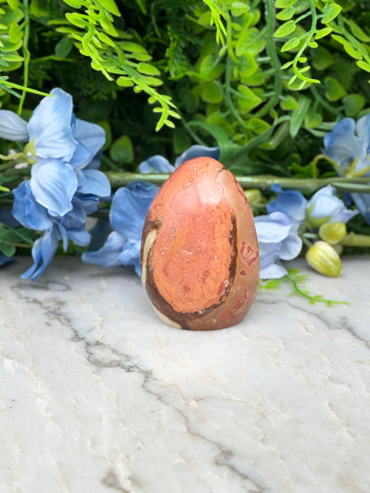 a brown and orange stone, possibly a jade egg, placed on a marble surface surrounded by blue flowers.