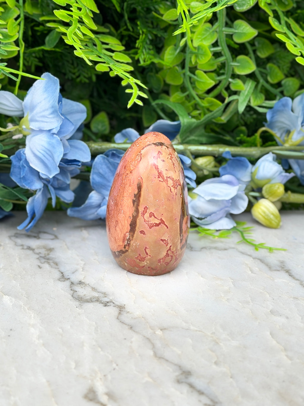 a pinkish-brown jade egg placed on a marble surface, surrounded by blue flowers and green foliage.