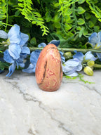 a pinkish-brown jade egg placed on a marble surface, surrounded by blue flowers and green foliage.